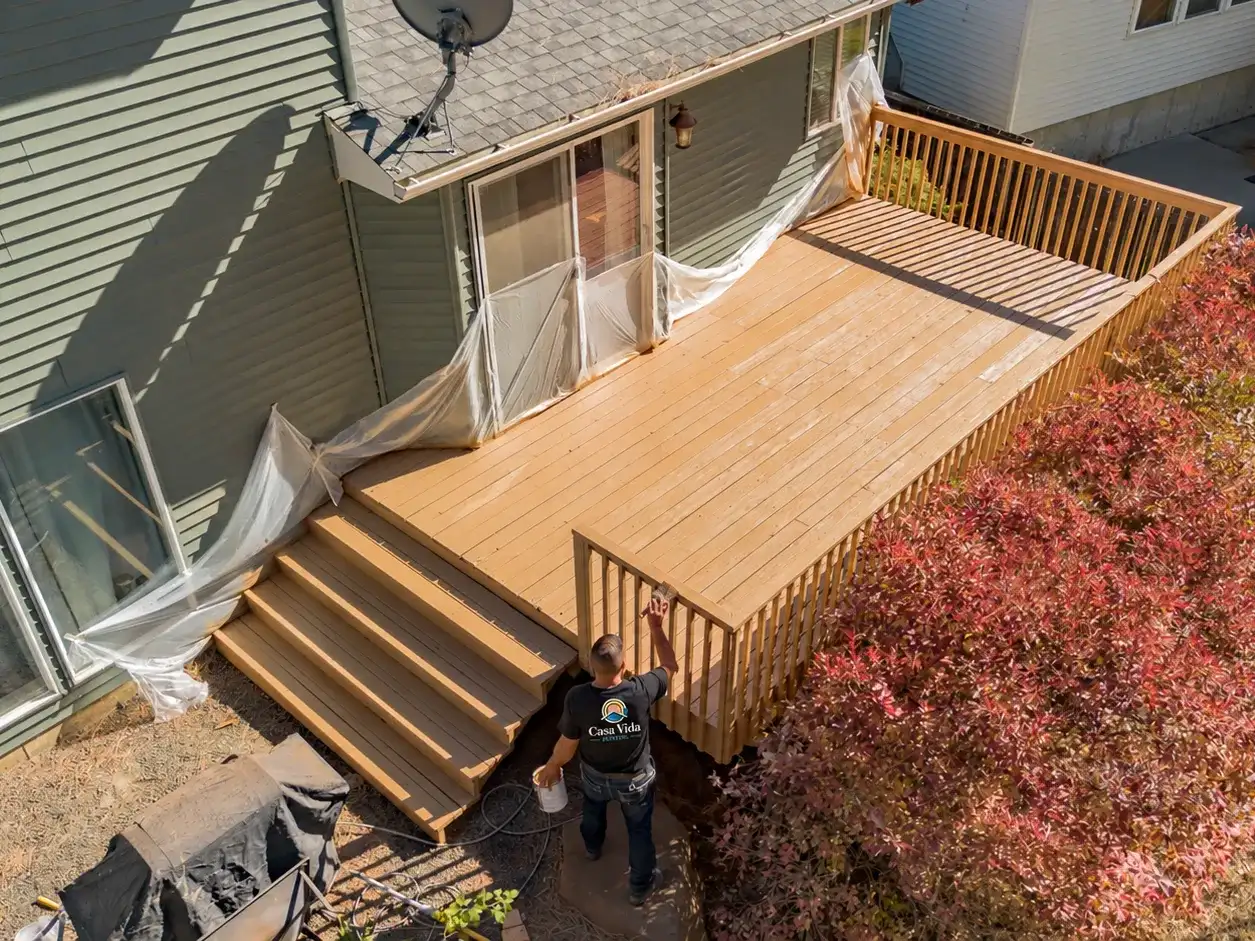 Drone view of a whole backyard deck and its stairs, freshly stained, with a Casa Vida painter staining the railing and the back windows masked in plastic