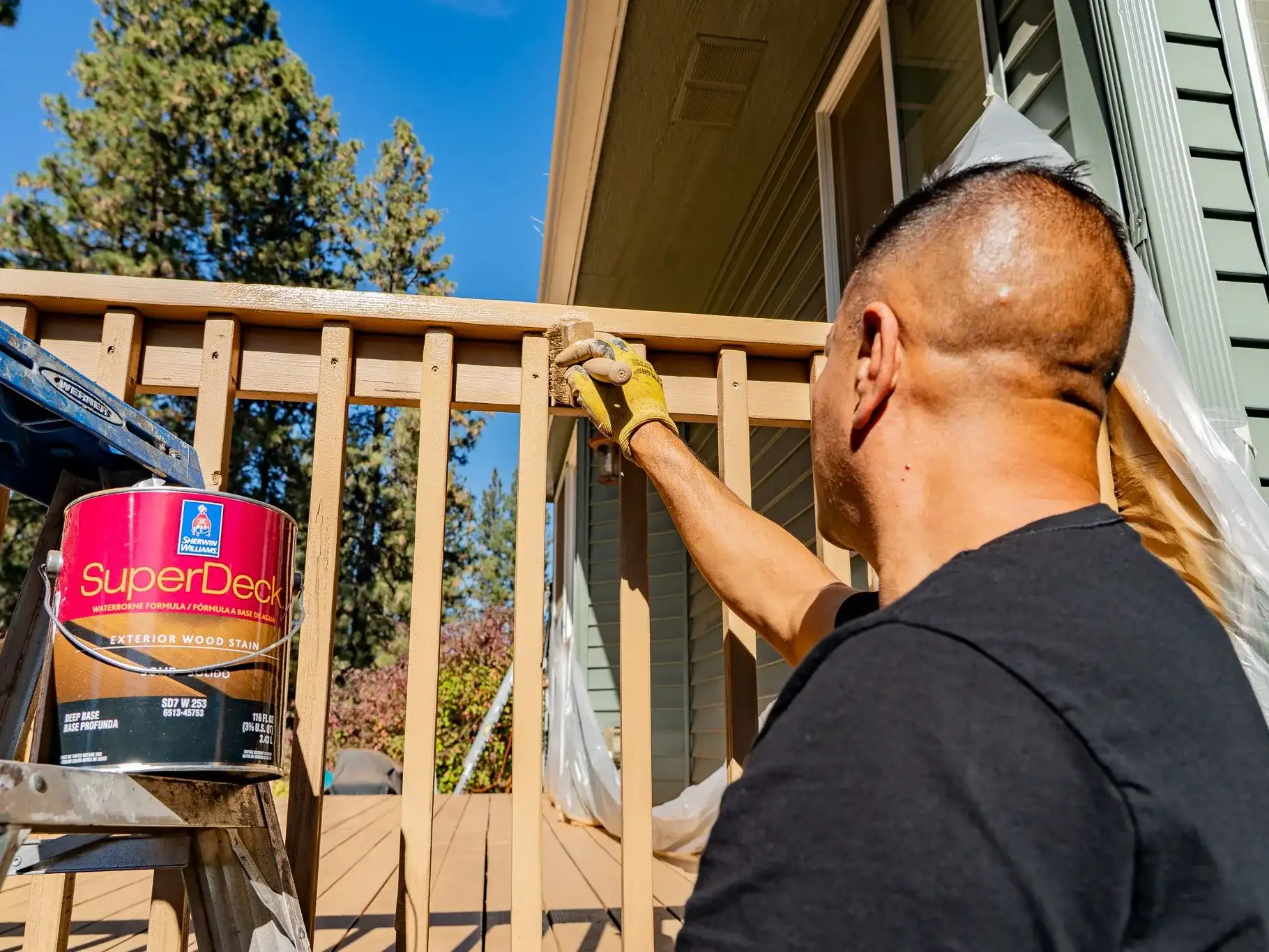 Casa Vida painter brushing stain onto a deck railing, a can of Sherwin-Williams SuperDeck exterior wood stain on the ladder