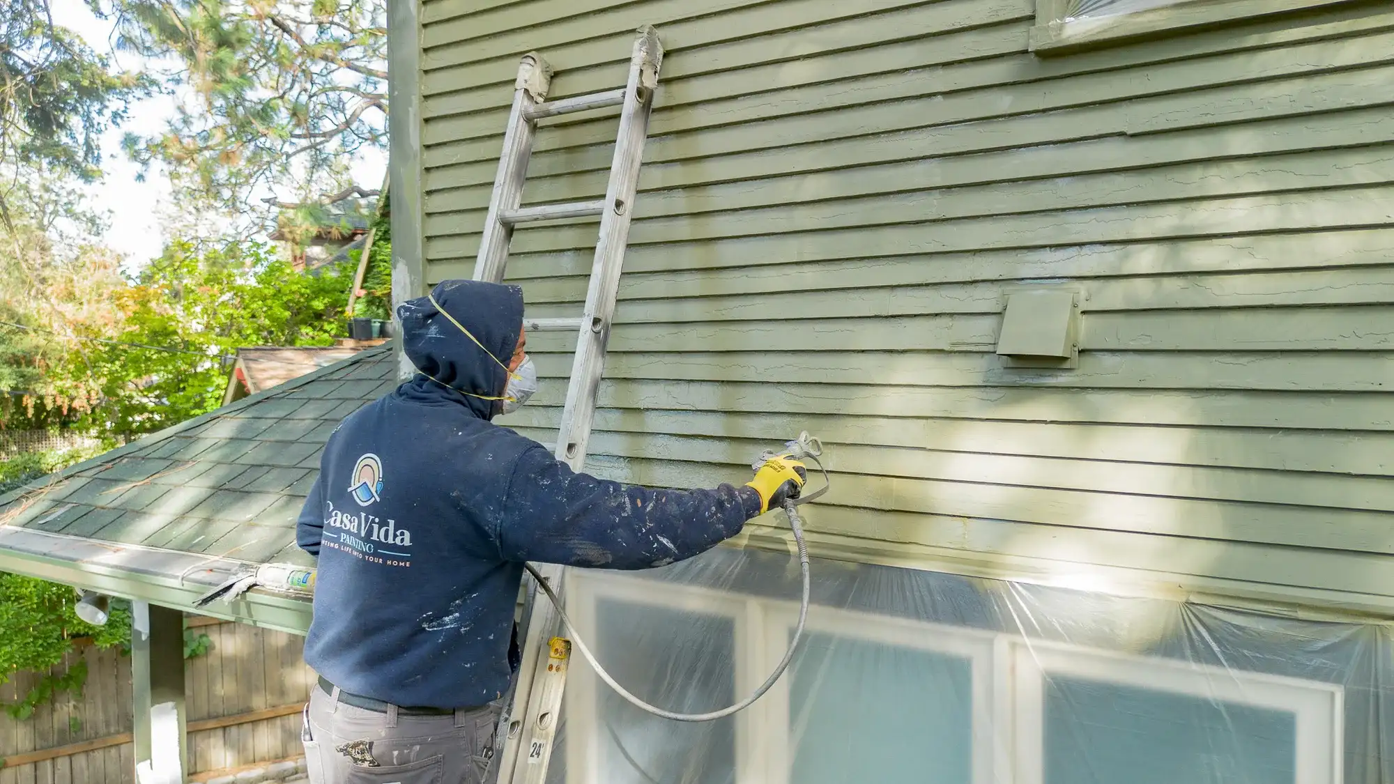 Casa Vida painter on a ladder spraying green siding, with the windows below masked in plastic