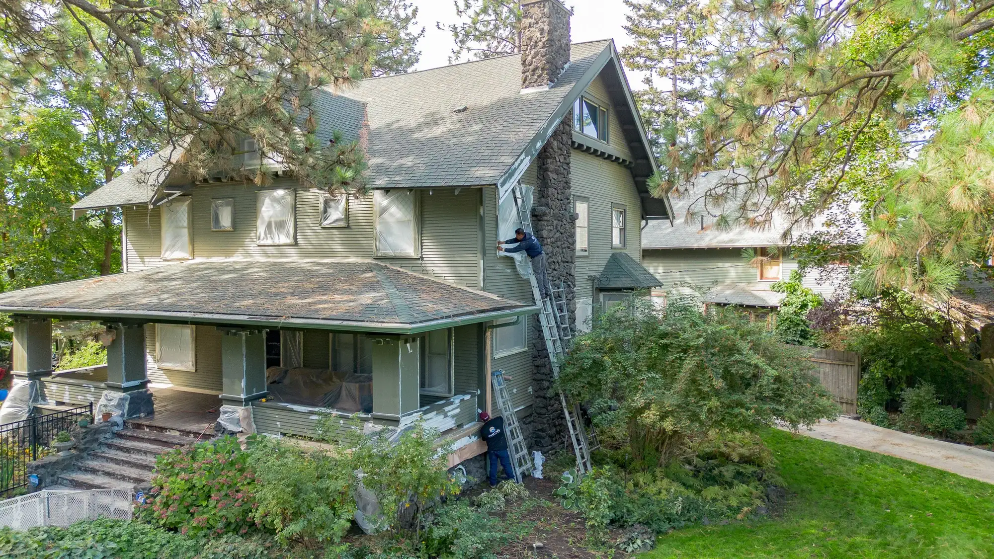 Drone view of a green Craftsman home in Spokane with the windows masked and Casa Vida painters on ladders