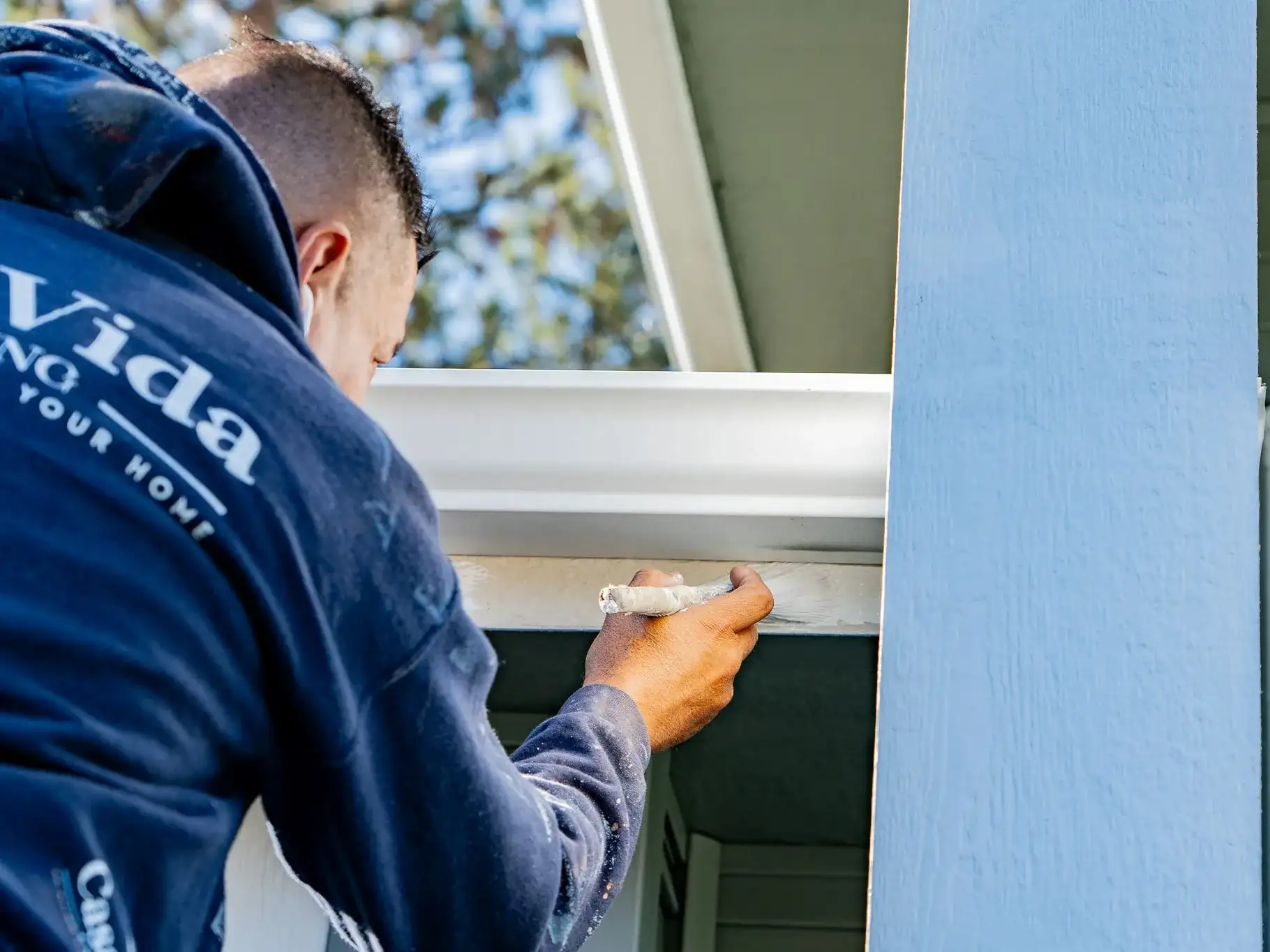 Casa Vida painter in a company hoodie brushing white paint onto trim under the eaves