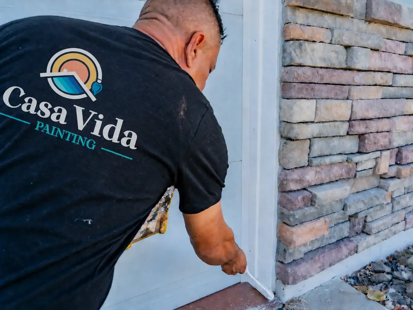Casa Vida painter, the company logo on the back of his shirt, brushing the base of the white trim in a clean line along the stone