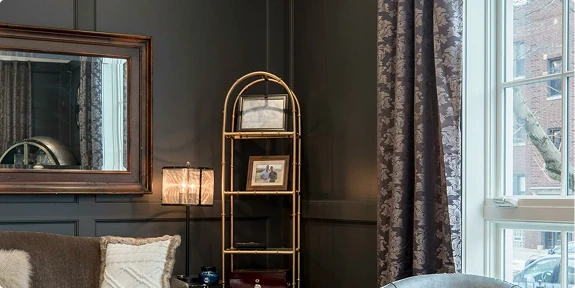Elegant living room corner featuring dark charcoal walls, a large window with patterned curtains, a gold-framed shelving unit displaying framed photos and decor, a table lamp, wall mirror, and part of a sofa with decorative pillows. Natural light streams through the window, highlighting the refined interior design.