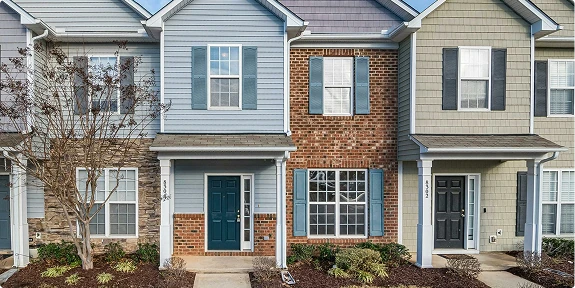 Row of modern attached townhomes with blue, gray, and brick exteriors, each featuring a covered front entry, large windows with shutters, landscaped planting beds, and paved walkways leading to the front doors.