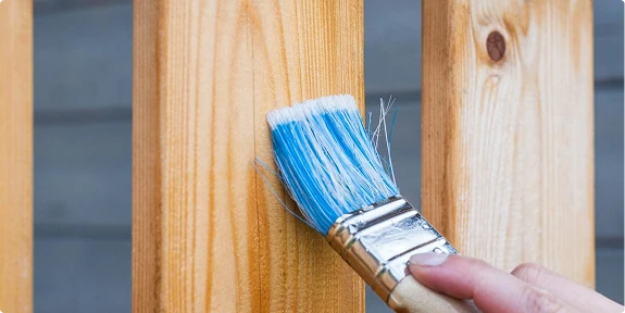 Close-up of a hand using a blue-bristled paintbrush to apply a fresh coat of clear finish or paint to a vertical wooden fence or railing, highlighting the natural wood grain.