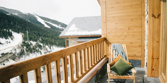 Wooden balcony with railing and outdoor chair overlooking a snow-covered mountain landscape. The cozy exterior space is attached to a timber-clad home and offers scenic views of alpine slopes and evergreen forests in winter.