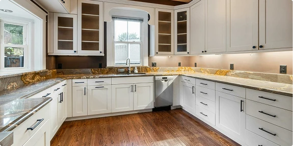 Bright, newly remodeled kitchen with white shaker cabinets, glass-front upper cabinets, granite countertops, stainless steel dishwasher, under-cabinet lighting, and hardwood floors. A large window above the sink brings in natural light, showcasing the clean, modern design.