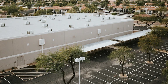 Aerial view of a large commercial building with a flat white roof, covered walkway entrances, and an expansive parking lot with marked spaces and mature trees. A surrounding suburban neighborhood is visible in the background.